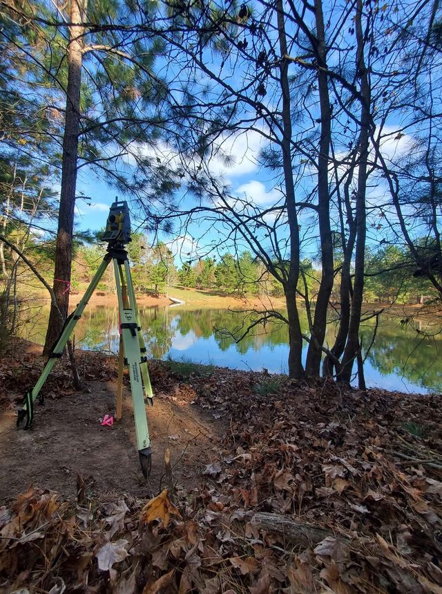 Total station on a tripod beside a pine-lined pond