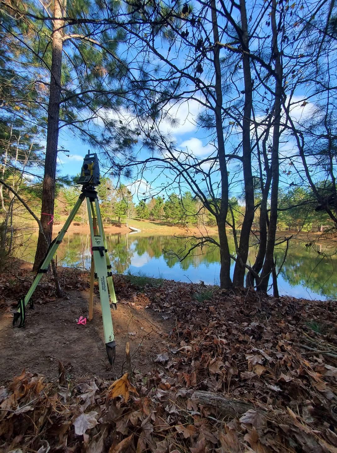 John Cowan & Associates total station on a tripod beside a pine-lined East Texas pond in the fall