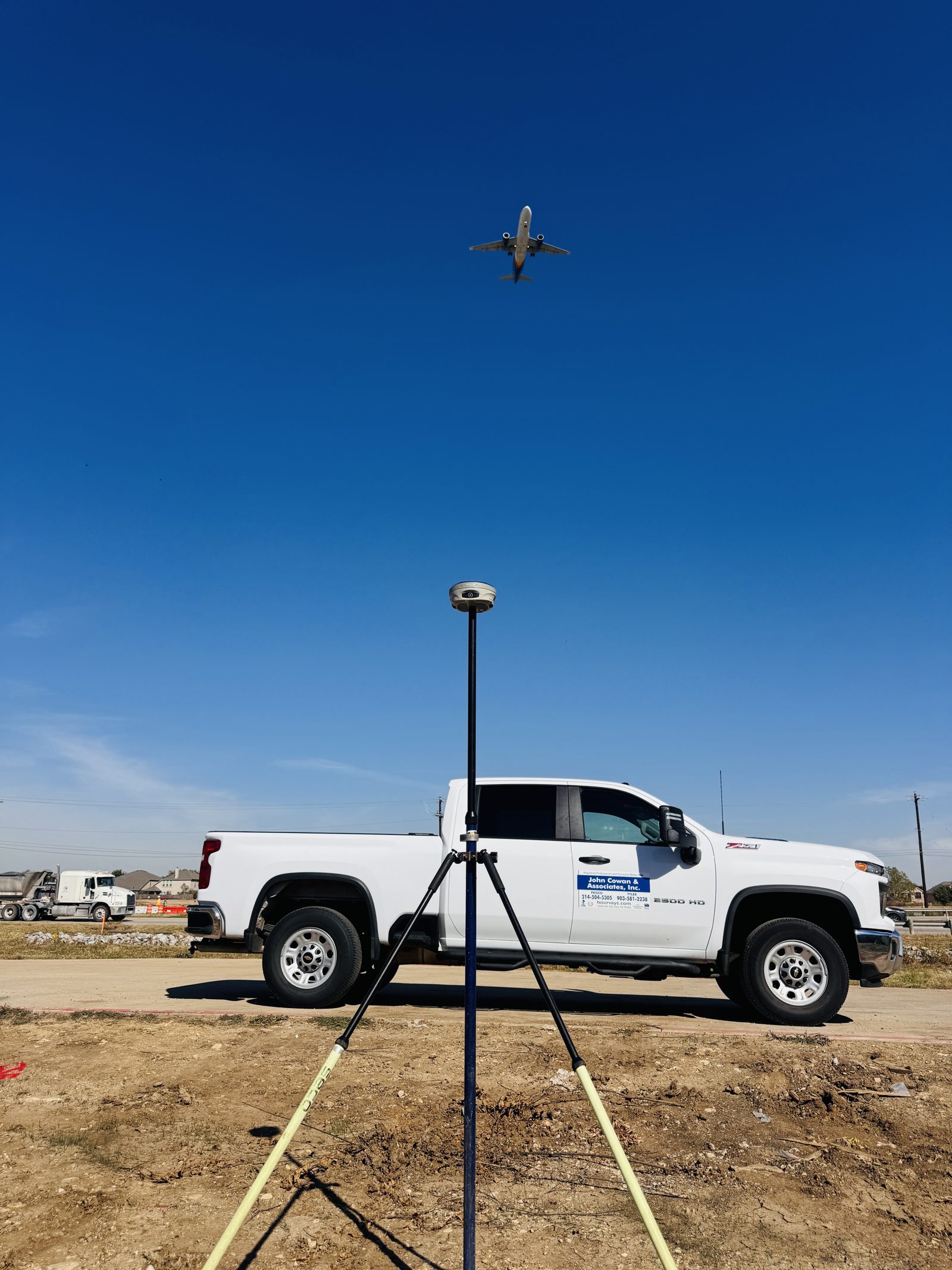 Carlson GNSS rover on a bipod-supported pole at a dirt job site, with a John Cowan & Associates truck behind it and a jet overhead