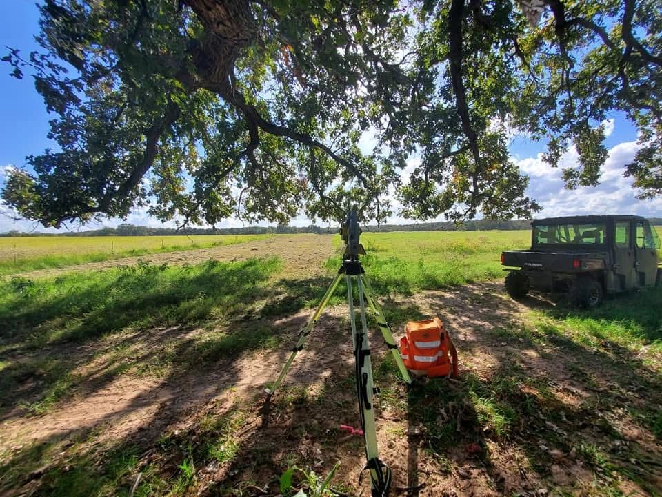 John Cowan & Associates total station set up under a large oak in an East Texas pasture, with the crew UTV nearby